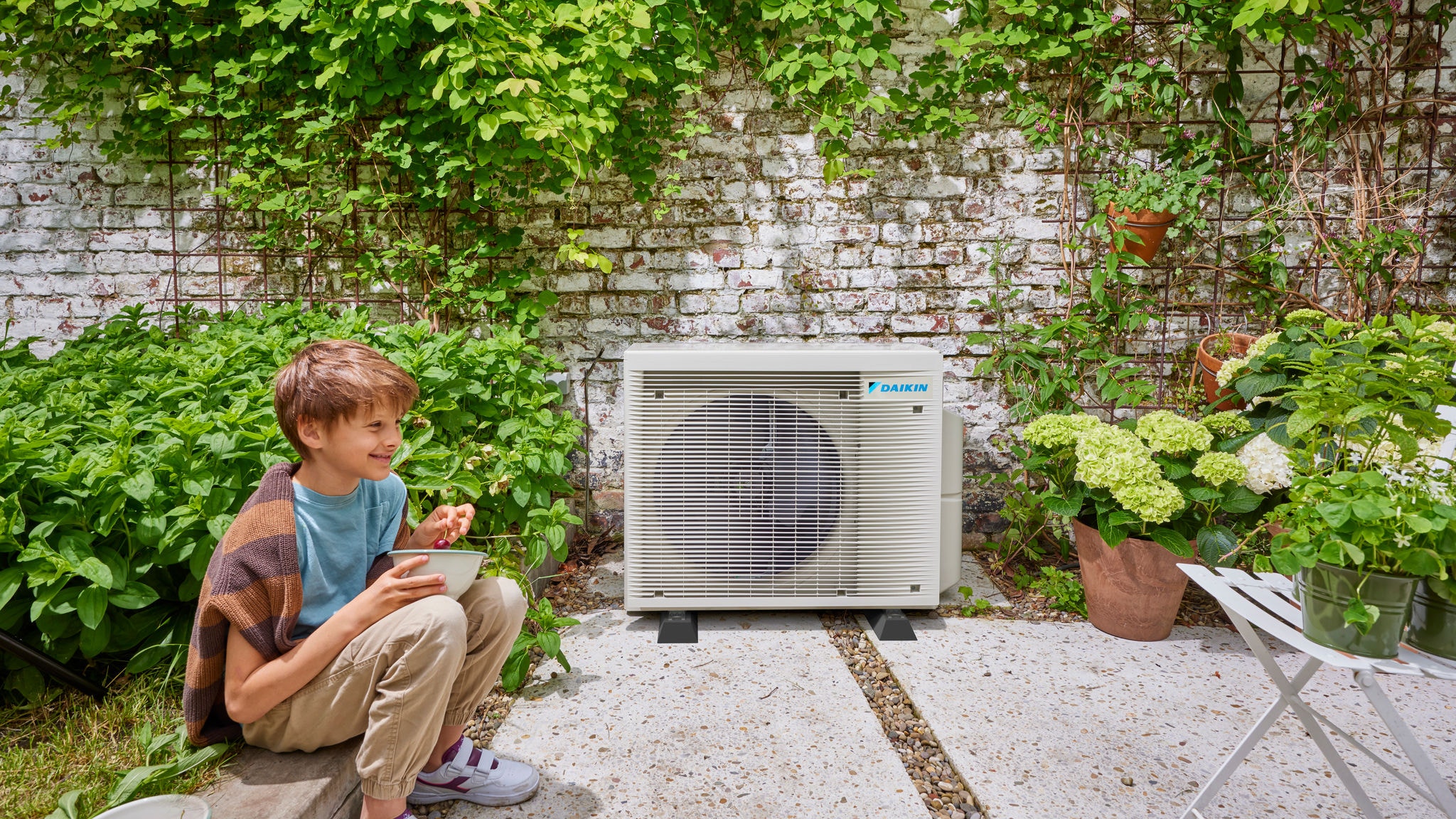 Un niño cerca de la unidad exterior de un equipo de aerotermia híbrido Daikin, que combina la eficiencia energética con un bonito jardín
