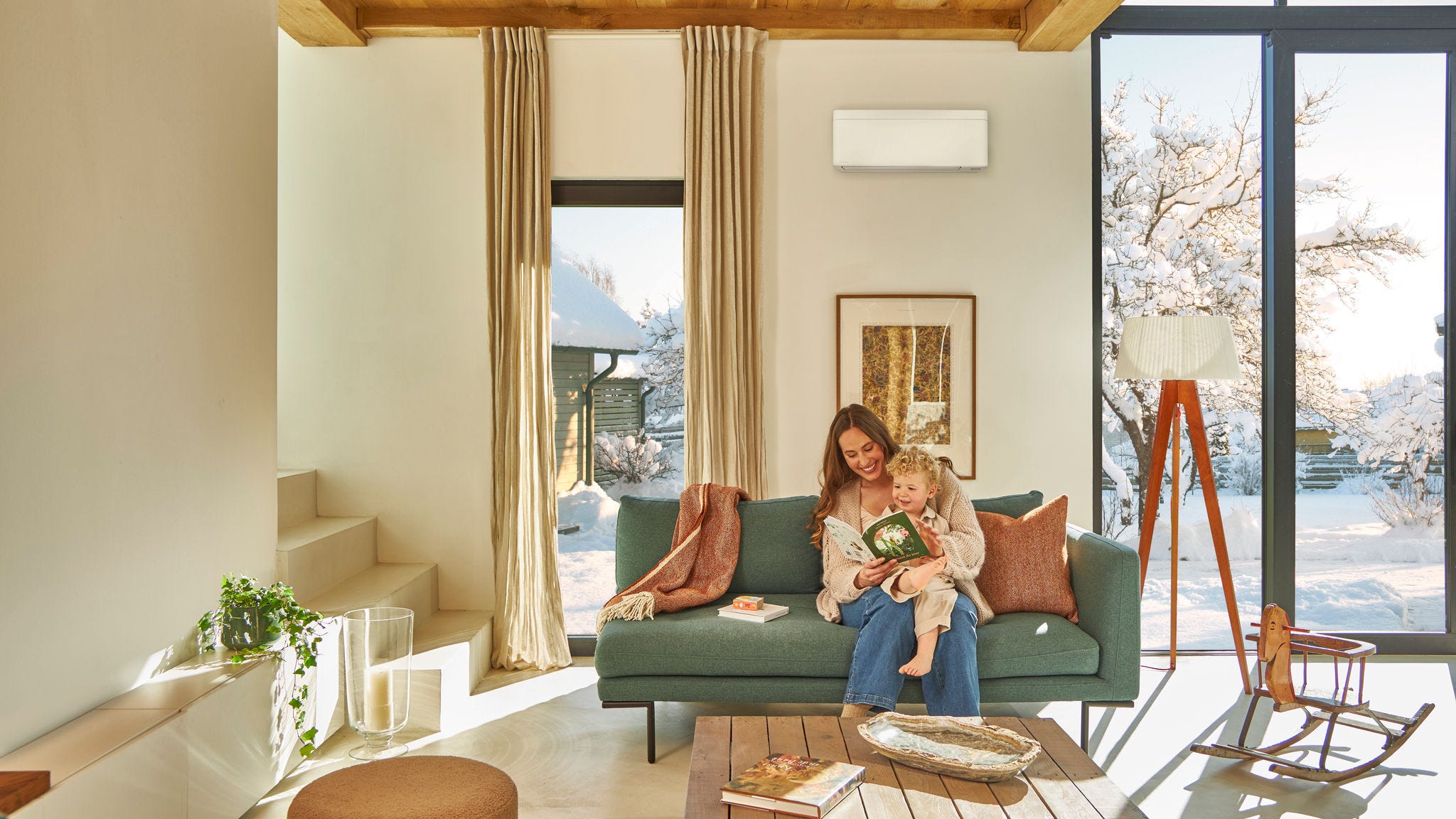 Madre sentada en el sofá leyendo un libro infantil con su niño pequeño en el regazo y disfrutando de un entrono cálido mientras nieva fuera gracias al sistema de aerotermia
